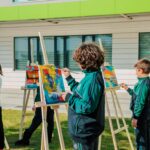 Children painting colorful designs on easels outdoors in school courtyard.